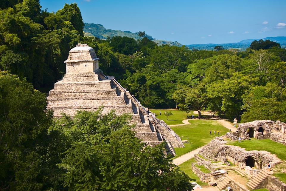 A multi-tiered, pyramidical stone temple surrounded by a dense forest with some visitors at the foot of the building.