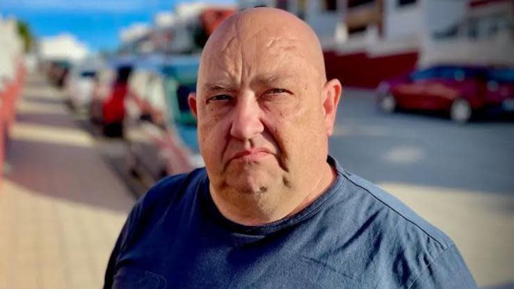 Billy George stands in the middle of a street looking at the camera. He is bald and is wearing a navy blue t-shirt. The sun is shining creating shadows from the cars and houses which line both sides of the street behind him. The background is blurred.