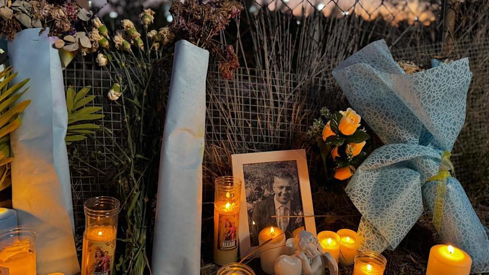 A small black and white photograph of John George has been placed in the grass beside a wire fence. Candles and flowers surround it lighting up the fence and surrounding grass. John is dressed in a suit in the photograph and is smiling. Beyond the fence a small amount of orange light fills the night sky. 