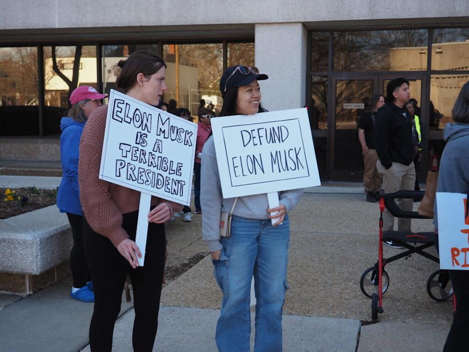 Protesters hold signs reading “Elon Musk is a terrible president” and “Defund Elon Musk.”