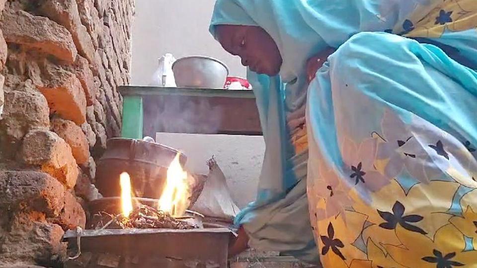 Manahel is crouching down by a fire, with cooking pots in the background. Her robe and headscarf are pale blue with yellow and dark brown flowers.  