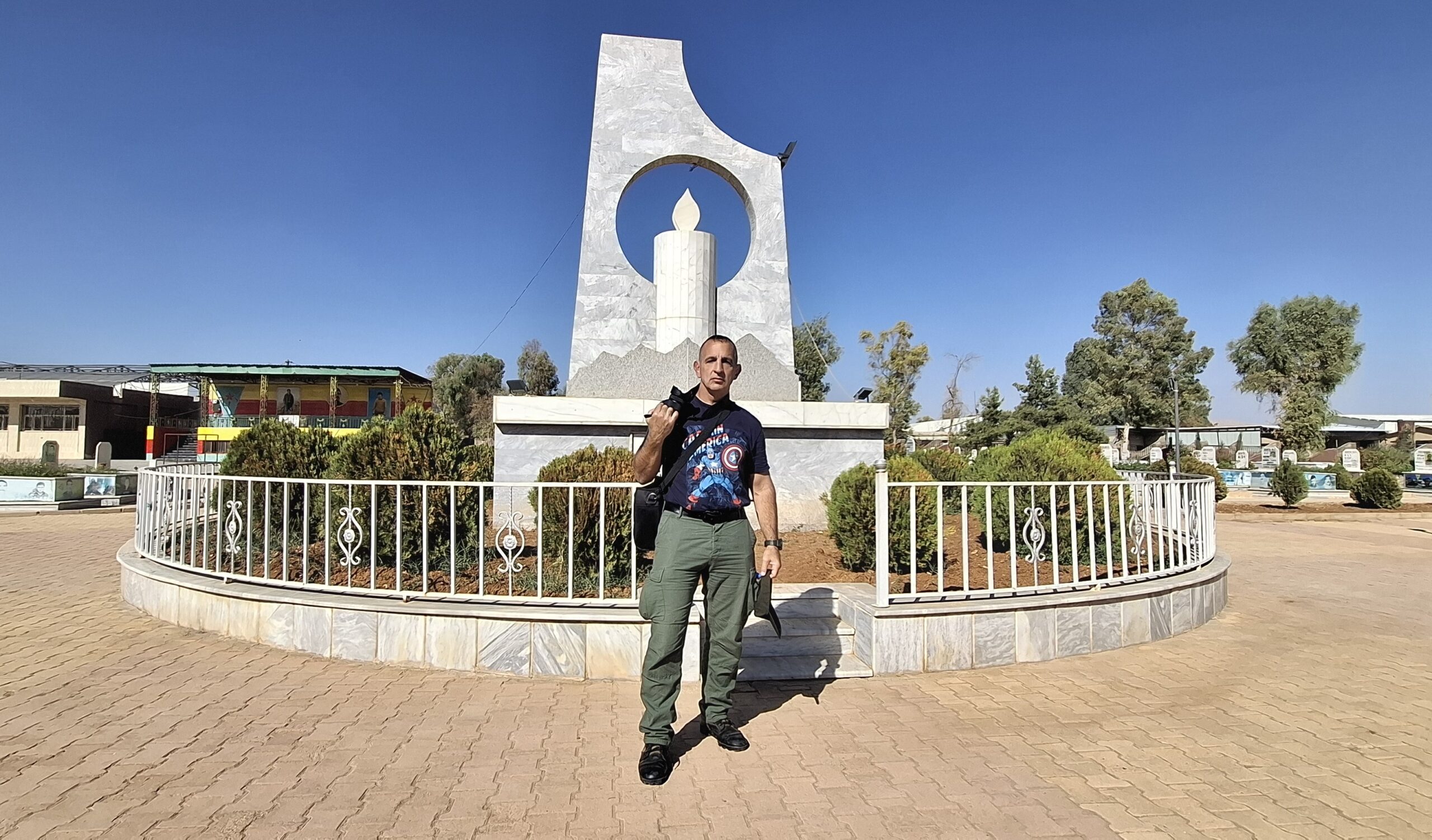 Man standing in front of a monument with a candle design, surrounded by greenery and buildings under a clear blue sky.