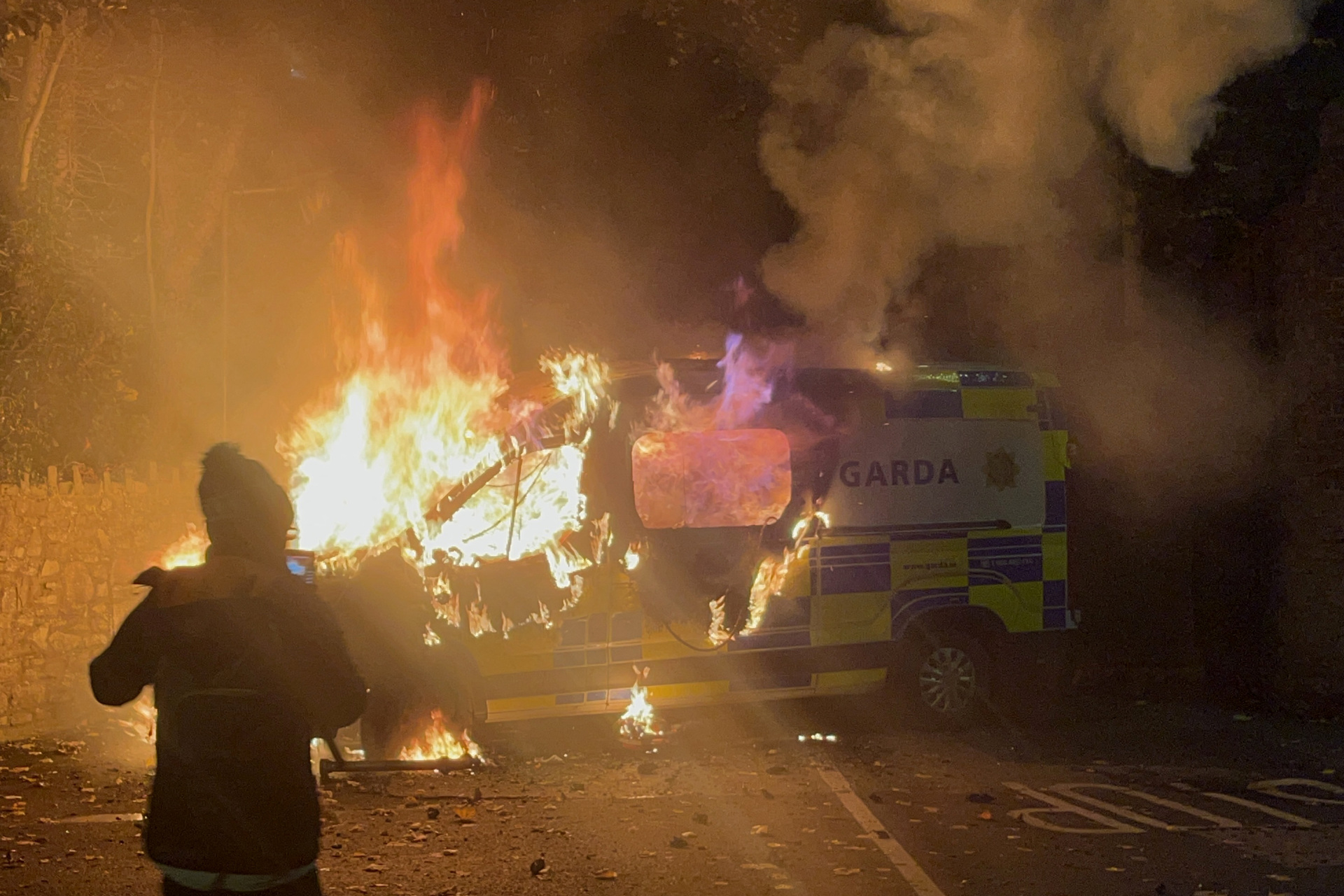 Onlookers film a police vehicle burning as a demonstration outside a hotel housing asylum seekers turns violent in Saggart, south-west of Dublin in Ireland on October 21, 2025. Police in riot gear clashed with at least 500 protesters in Dublin on Tuesday outside an asylum seeker hotel, following allegations that a 10-year-old girl was sexually assaulted. (Photo by Peter MURPHY / AFP) (Photo by PETER MURPHY/AFP via Getty Images)