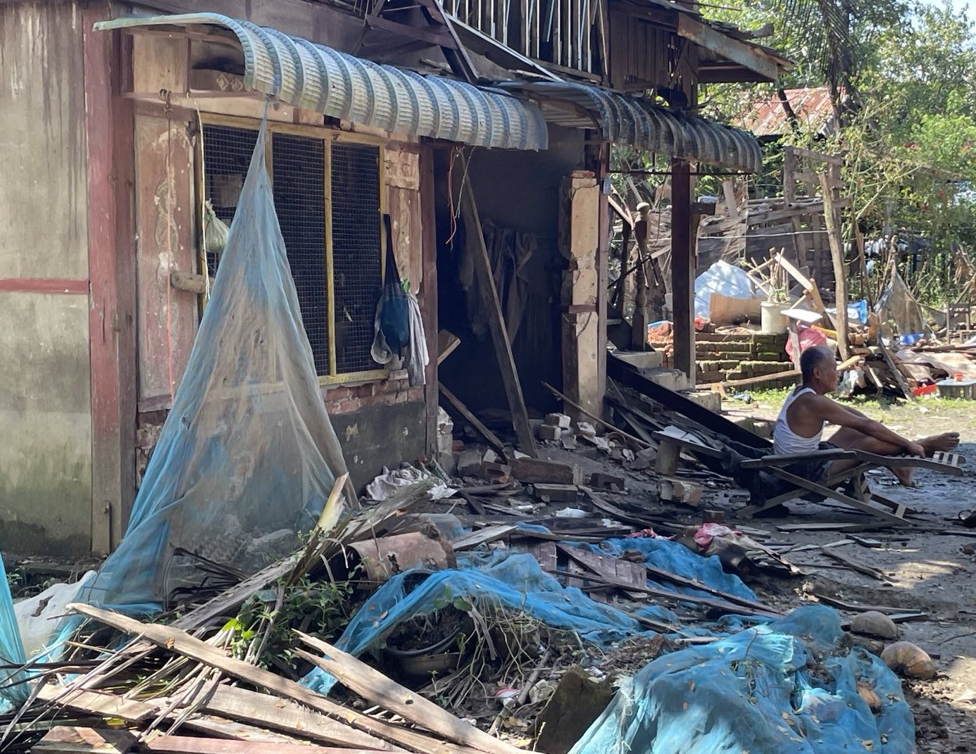 An elderly man sits amid debris and damaged structures, highlighting the impact of natural disasters on local communities.