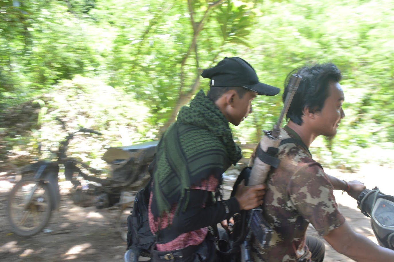 Two individuals riding a motorcycle through a dense, green landscape, one wearing a camouflage shirt and the other in a black cap and scarf.