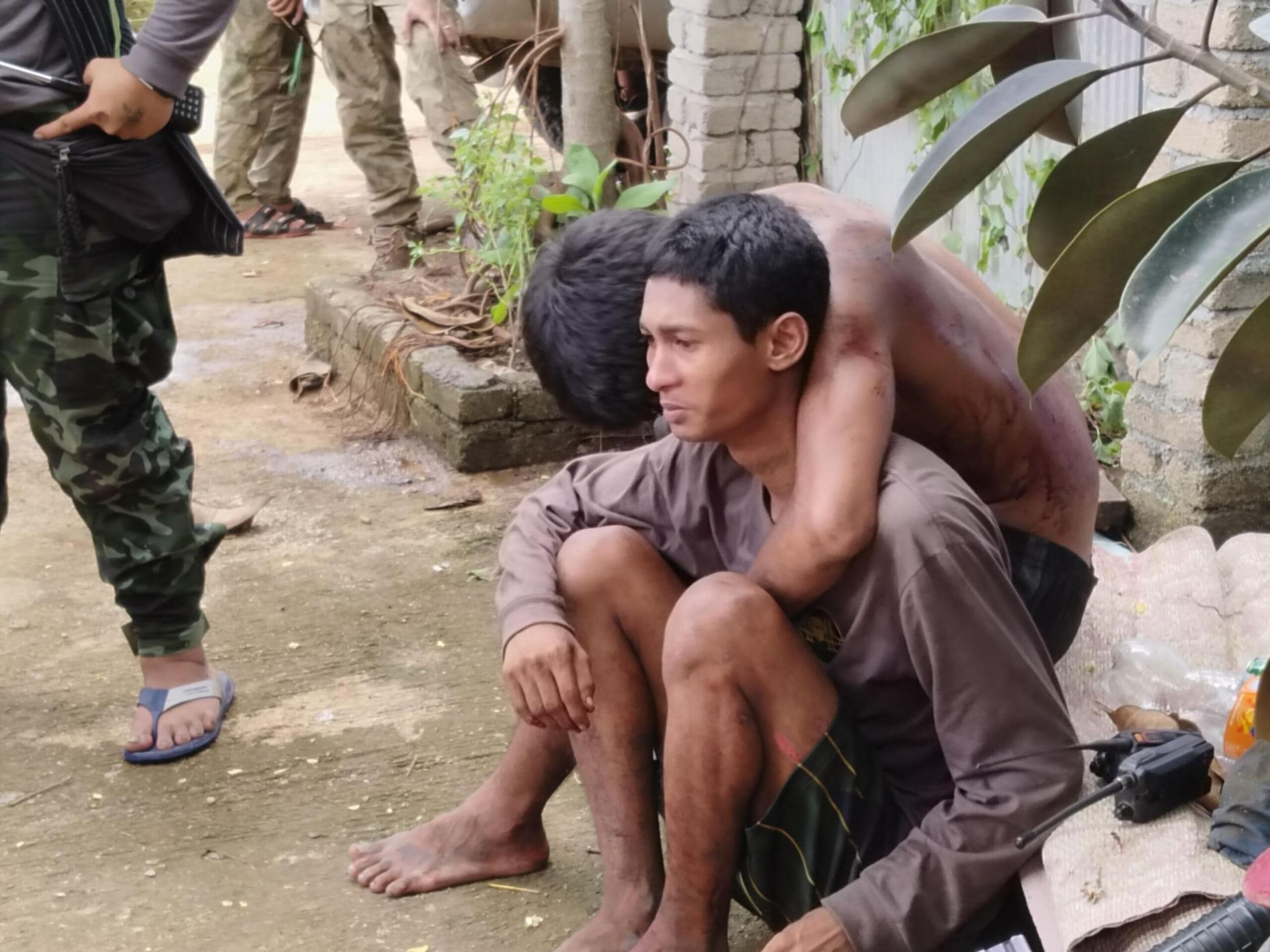 Two individuals sit on the ground, one appearing distressed and the other embracing him, with a group of people in the background.