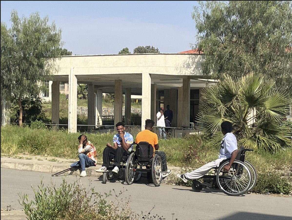 Group of individuals, including wheelchair users, socializing outside a building in a green, open area, showcasing community engagement and accessibility.