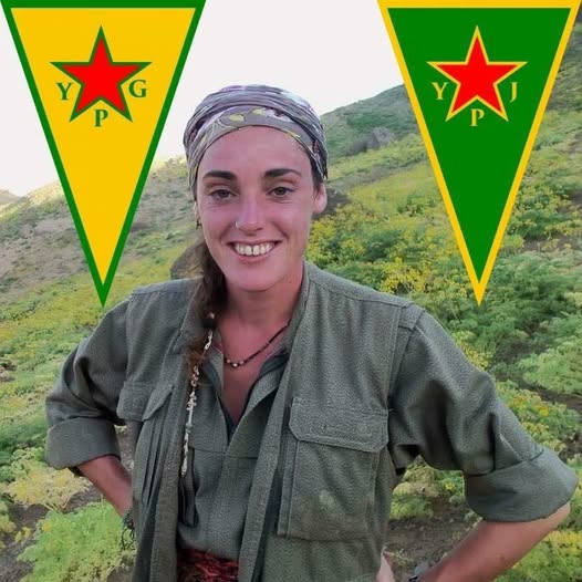 Smiling woman in military attire stands in a green landscape with YPG and YPJ flags in the background, symbolizing Kurdish women's resistance.