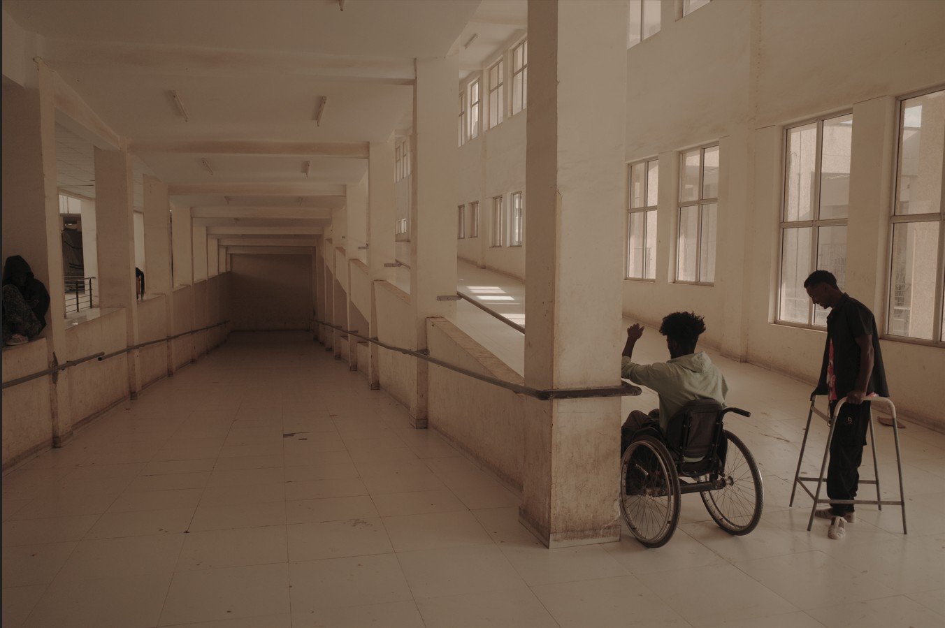 Two individuals navigate a spacious, well-lit corridor; one uses a wheelchair while the other walks with a walker, highlighting accessibility challenges.