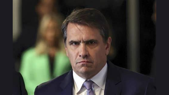 Serious-looking man in a suit with a focused expression, standing in a formal setting with blurred figures in the background.