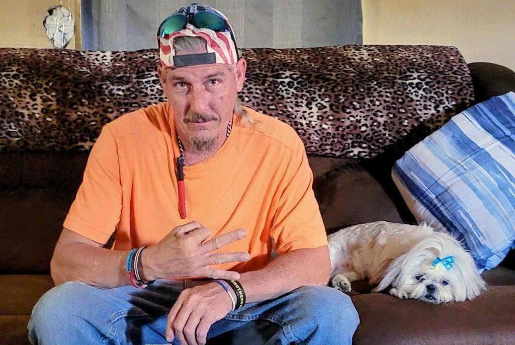 Man in an orange shirt making a hand sign sits on a couch with a small dog beside him, set against a leopard print background.