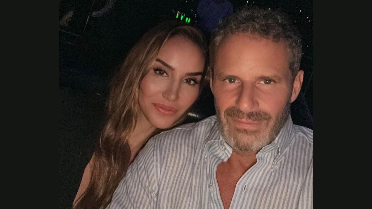 A couple poses for a selfie in a dimly lit setting, showcasing their smiles and stylish attire.