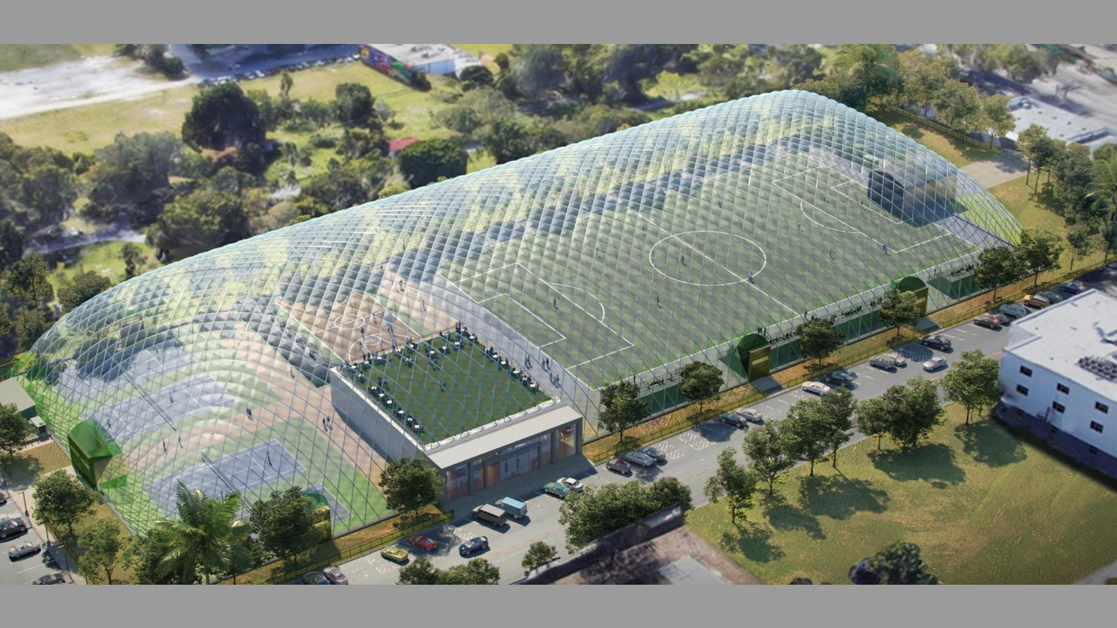 Aerial view of a modern indoor sports facility featuring multiple soccer fields under a large transparent dome, surrounded by greenery and parked vehicles.