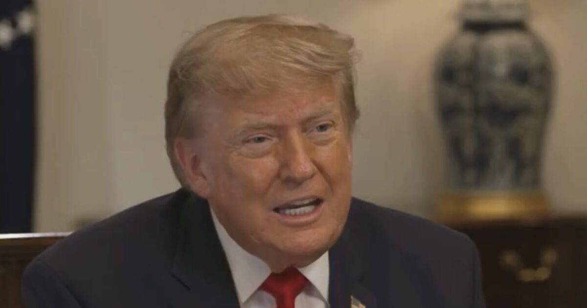 Donald Trump speaking during an interview, wearing a dark suit and red tie, with decorative background elements visible.