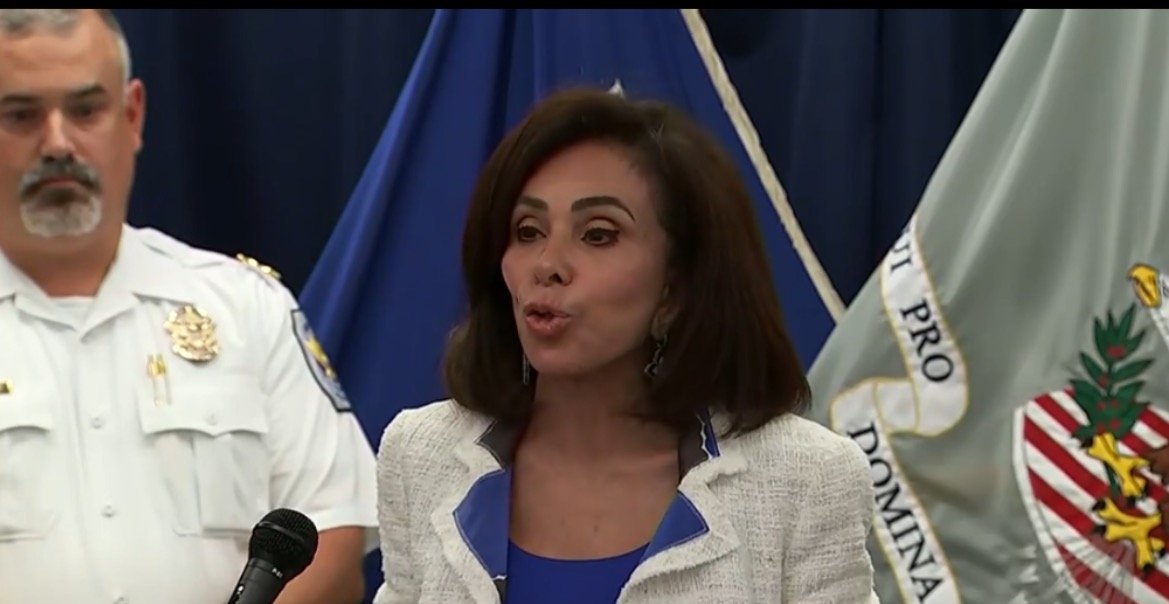 Official press conference featuring a woman speaking into a microphone, with a police officer in the background and flags displayed behind them.