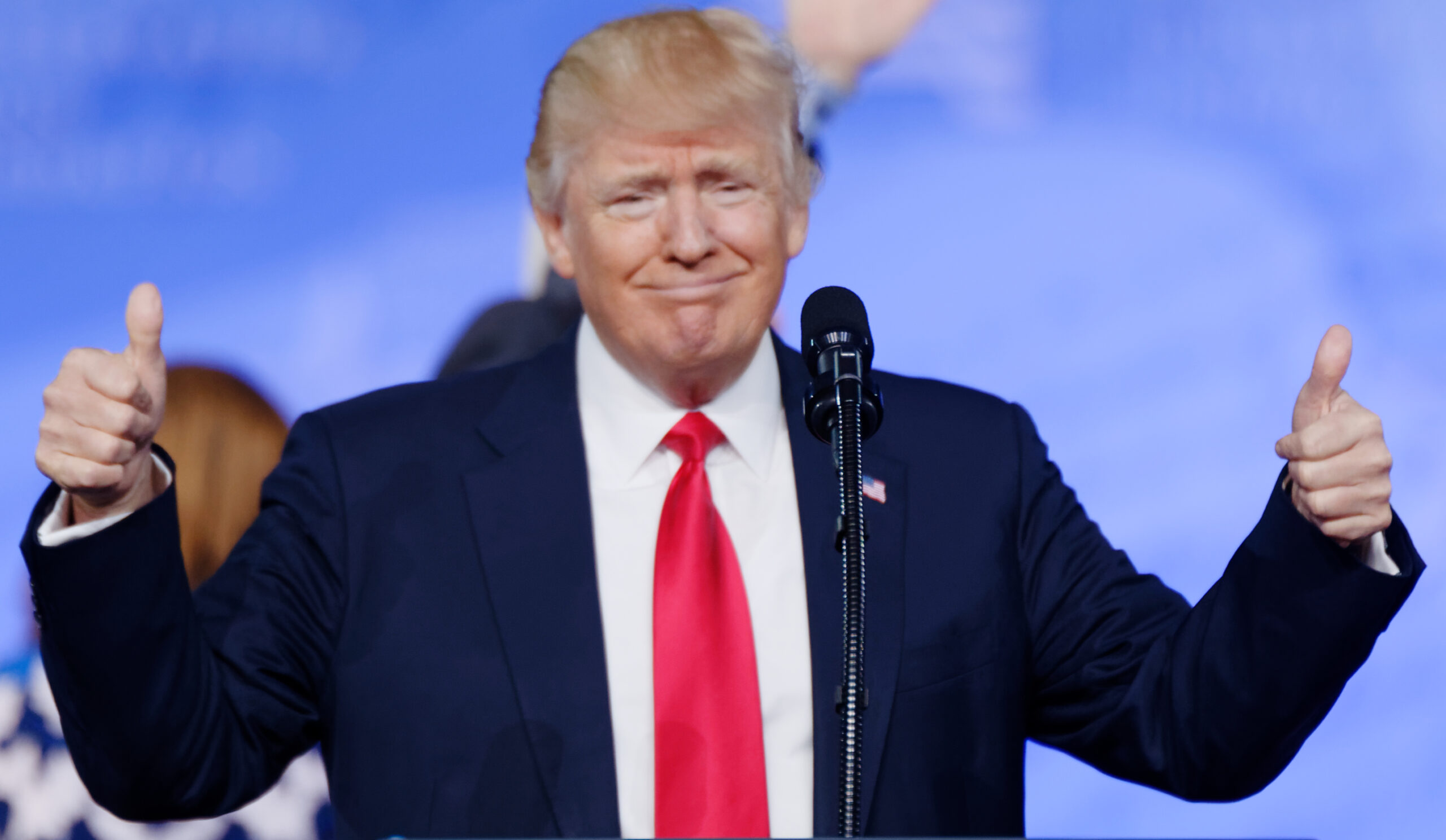 Donald Trump smiling and giving thumbs up while speaking at a podium during a public event.