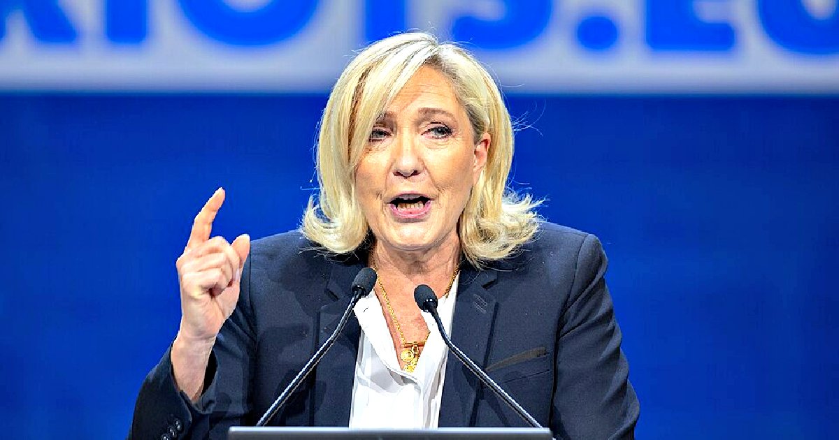 Politician delivering a speech with expressive hand gestures, wearing a black blazer against a blue background at a public event.