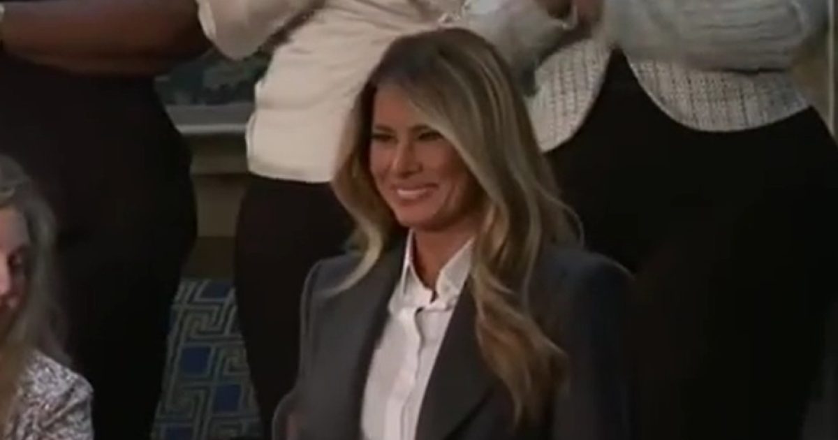 Melania Trump smiling while attending an event, surrounded by applauding audience members, wearing a stylish outfit with long hair.